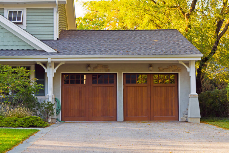 A suburban house with two wooden garage doors featuring garage door windows, grey siding, white trim, and a driveway. Trees with green leaves are visible in the background.