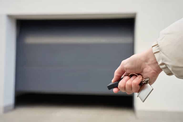 A person’s hand holding a remote and keys in front of a closed gray garage door, about to open it with a smart garage door opener.