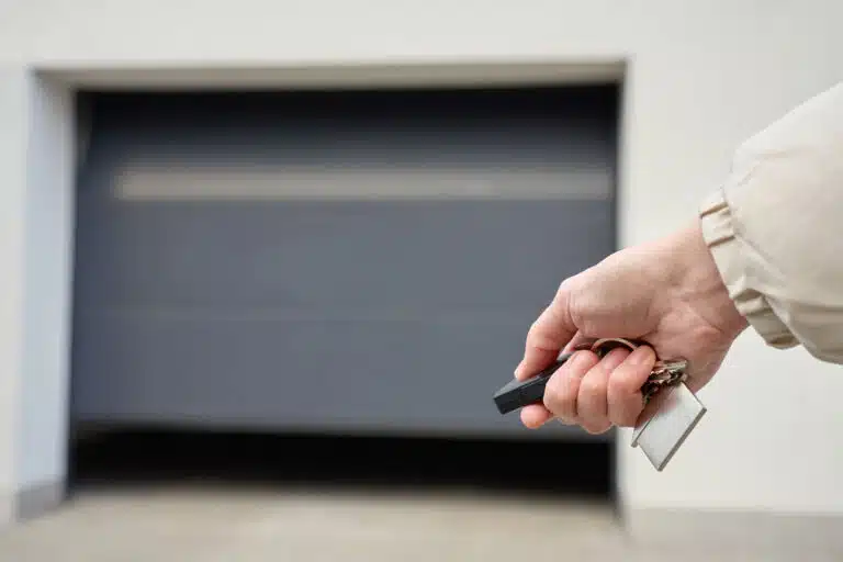 A person’s hand holding a remote and keys in front of a closed gray garage door, about to open it with a smart garage door opener.
