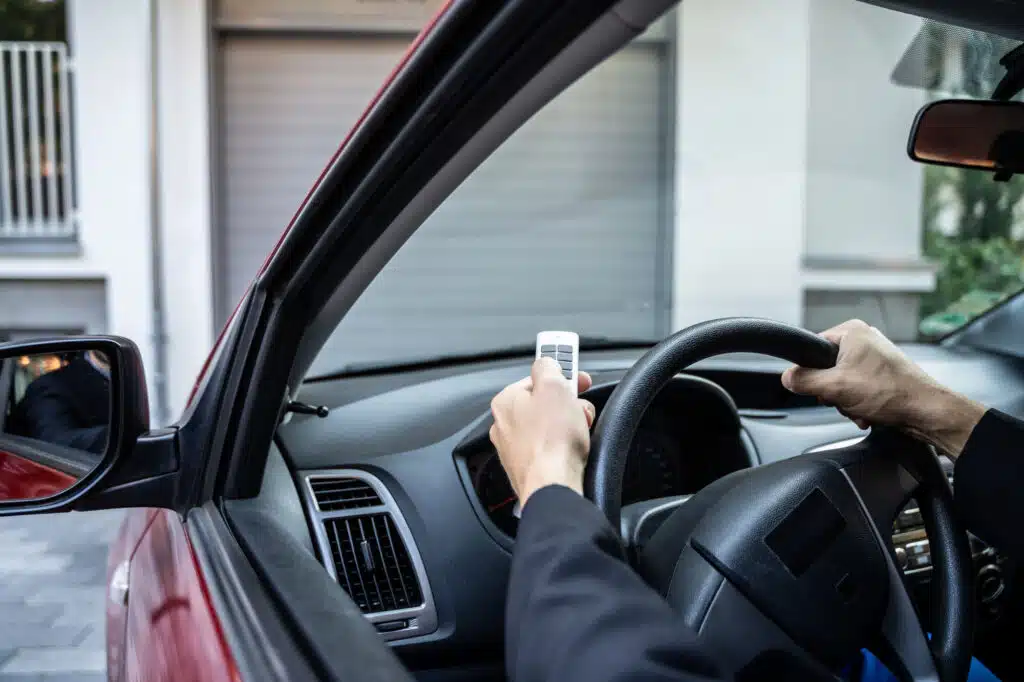 A person in a car uses a remote control to open a smart garage door opener. The driver’s hands are on the steering wheel, and the garage door is partially open in front of the red vehicle.